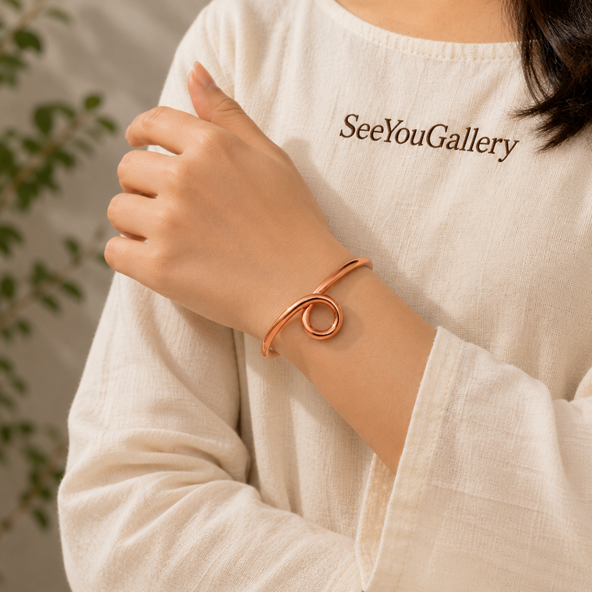 A person wearing a thin rose gold cuff bracelet with a double loop design, styled with a cream-colored linen top that has 'SeeYouGallery' printed on the chest.